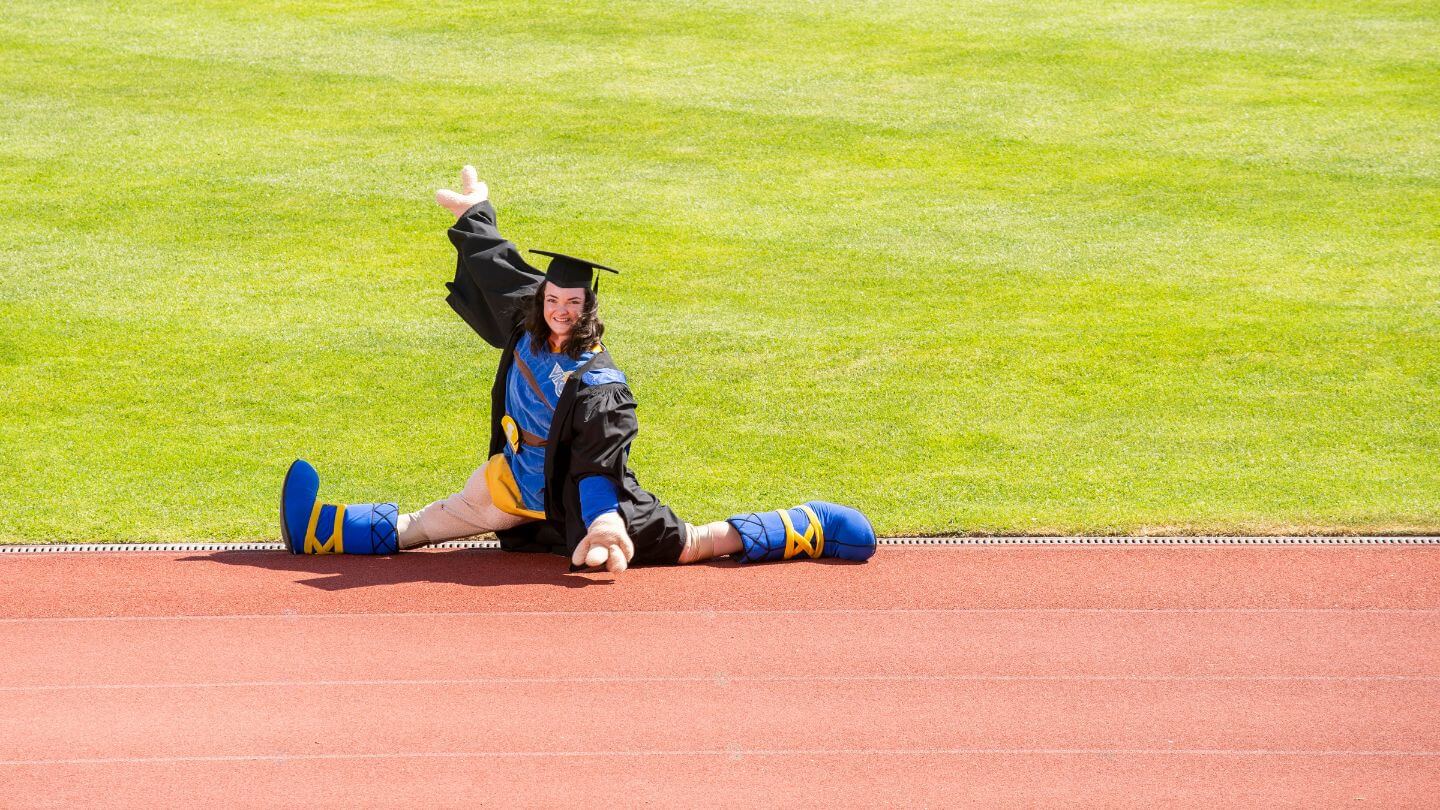 Women with dark hair in blue and yellow Norse God Thunder mascot costume and black graduation cap and gown performs the splits with a sports field in the background.
