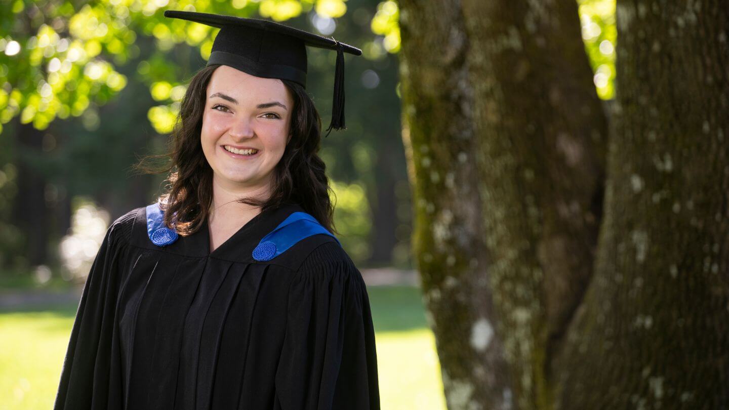 Woman with brown hair in black graduation cap and gown with royal blue smiling beside a tree