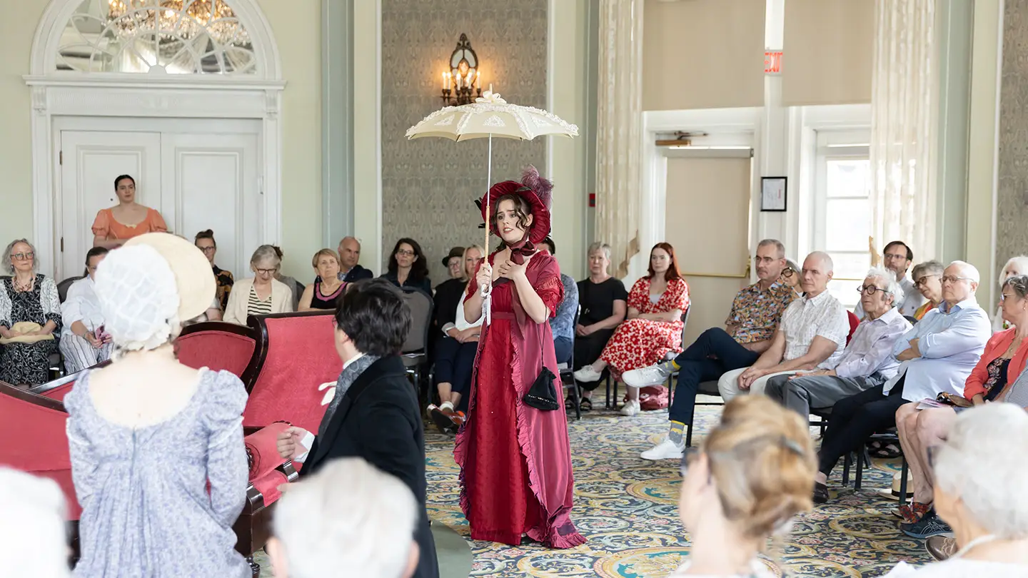 A singer holding a parasol sings in the middle of the room, surrounded by a seated audience. 