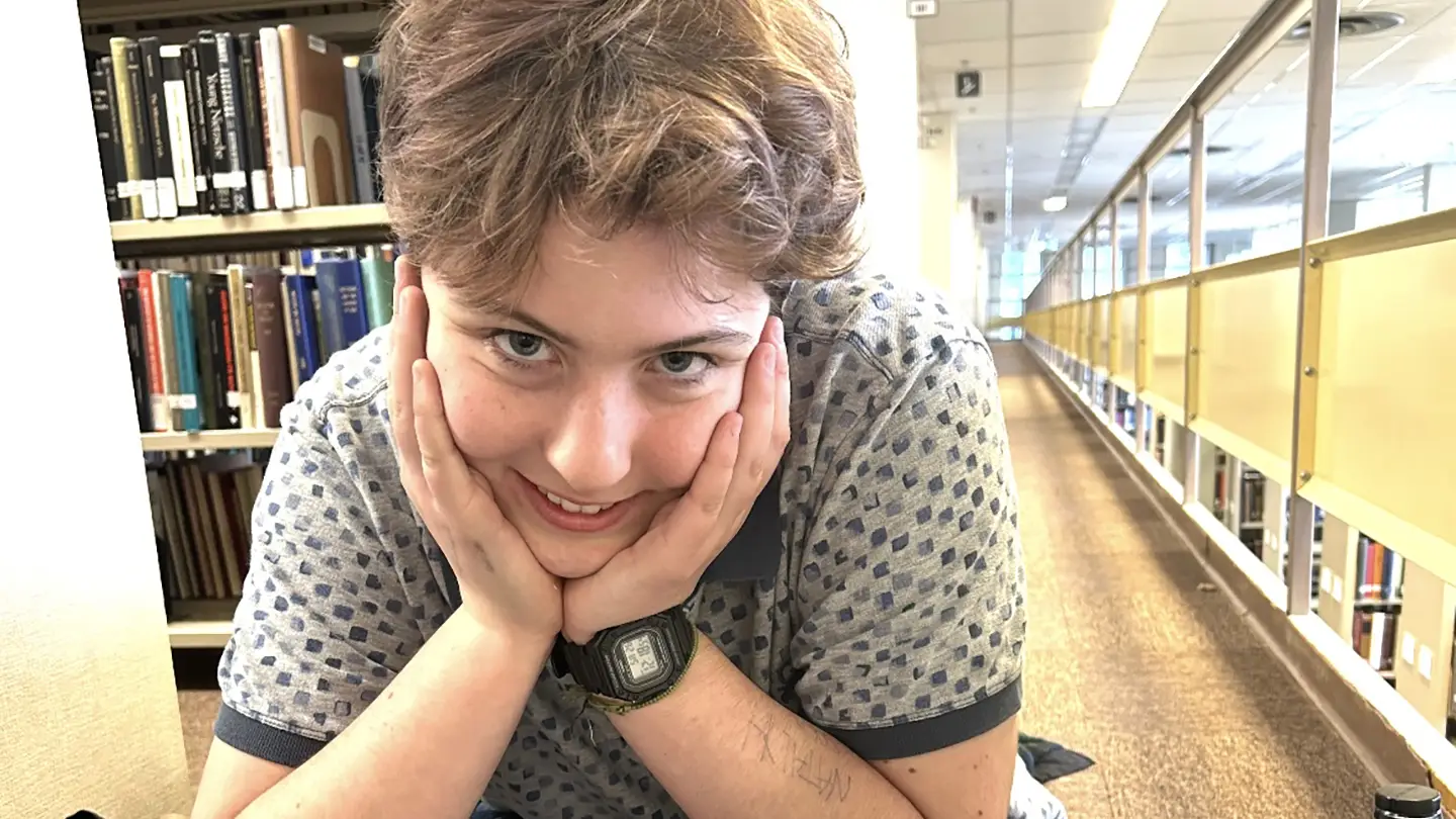 A student sits cross-legged on the floor of the library, in between rows of books. 