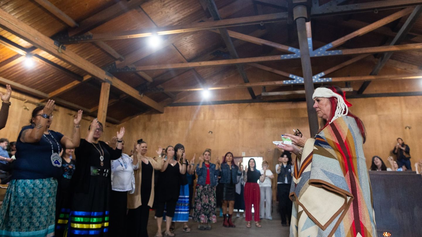 Dr. Robina Thomas stands wrapped in a ceremonial blanket surrounded by friends, family and colleagues who raise their hands in appreciation and respect.