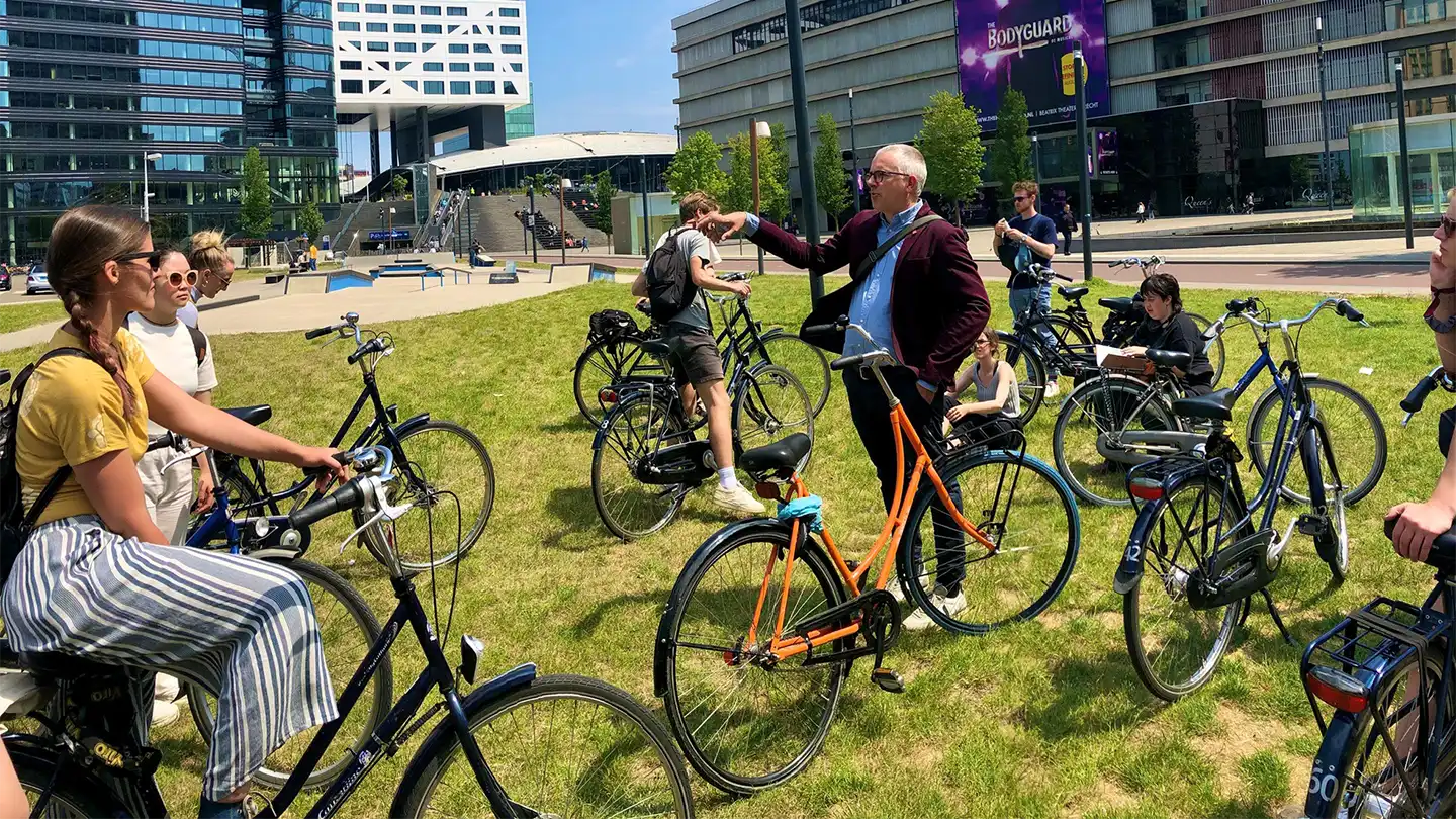 A group of people gather together in a grassy park area and stand with or on bicycles.