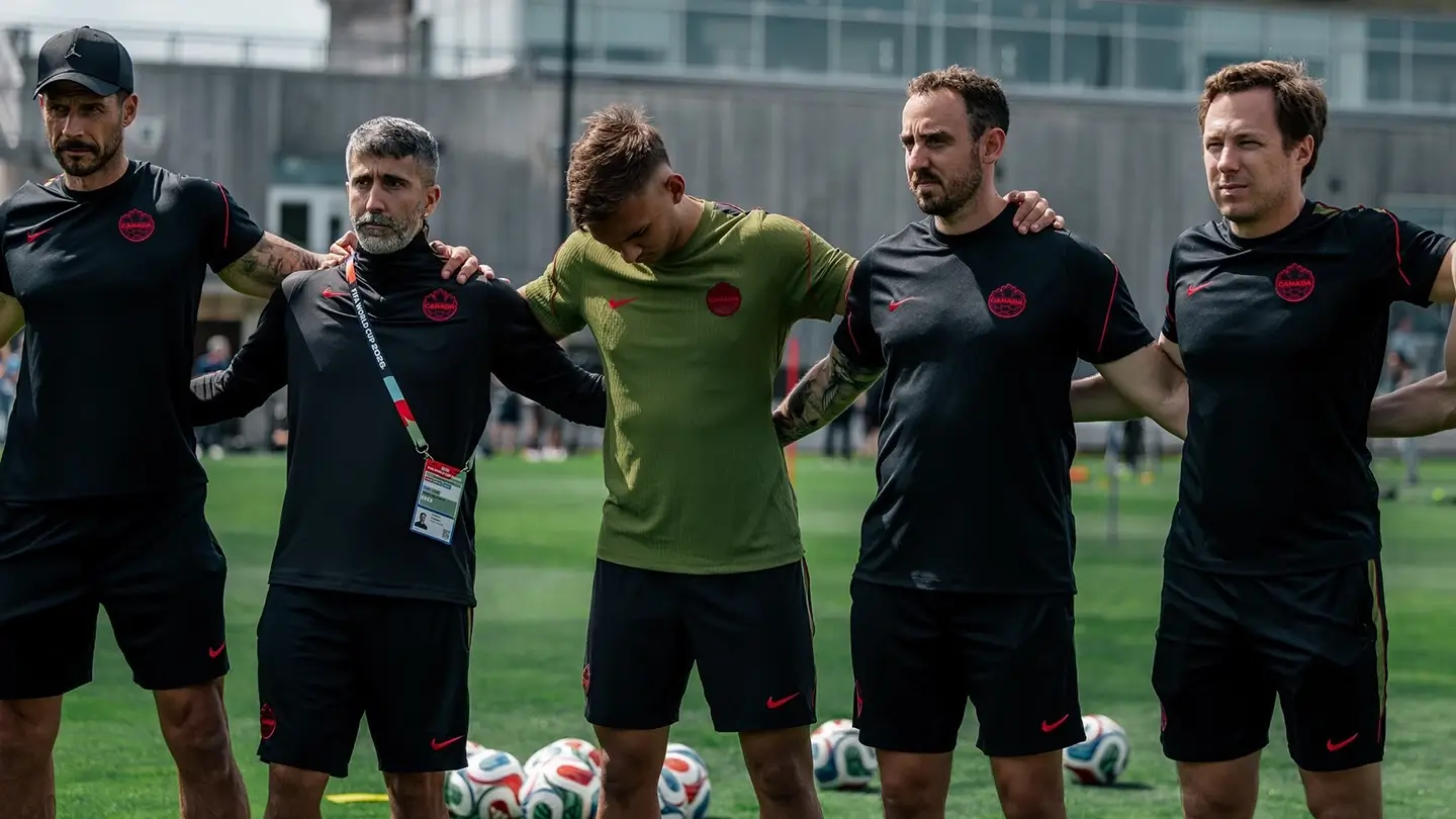 Five men stand together on a soccer field wearing team Canada soccer gear.