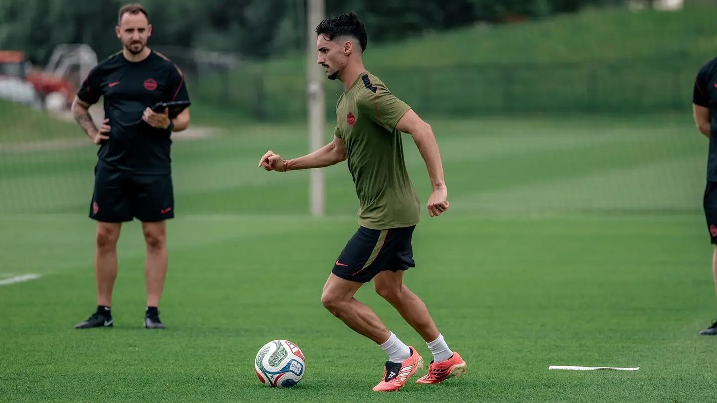 A soccer player prepares to kick a soccer ball on the field as Pat Cormier observes in the background.