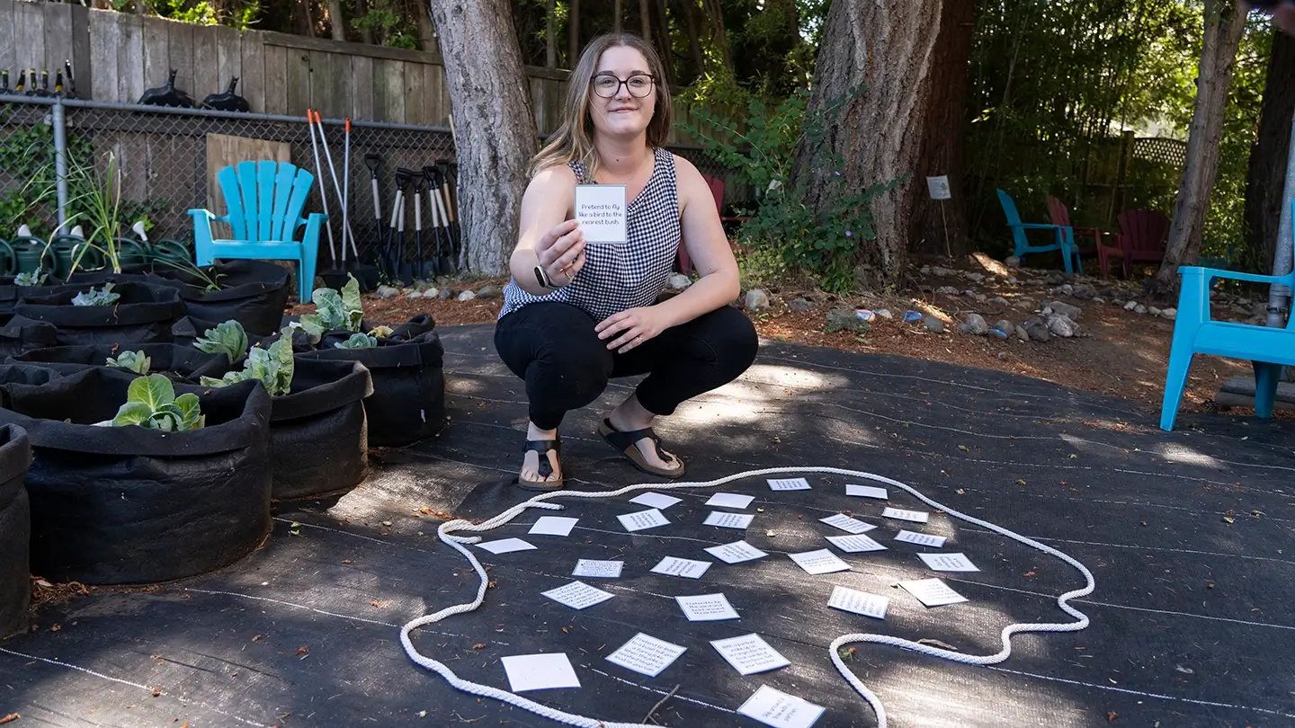 Merah Gasmo crouches in a garden with a collection of suggested activities written on small slips of paper.