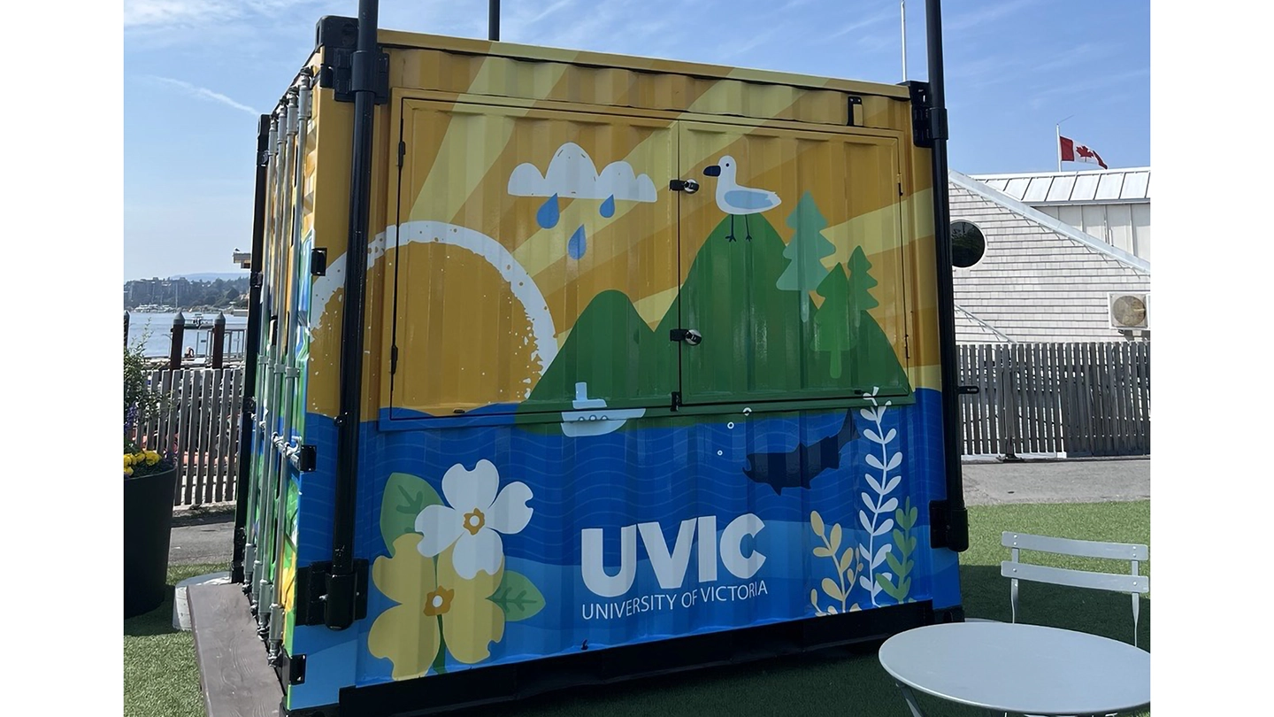 A shipping container vibrantly decorated with a sunset, mountains, birds, animals, sea life and plants found on Vancouver Island. The container is set up in Victoria's Ship Point harbour.