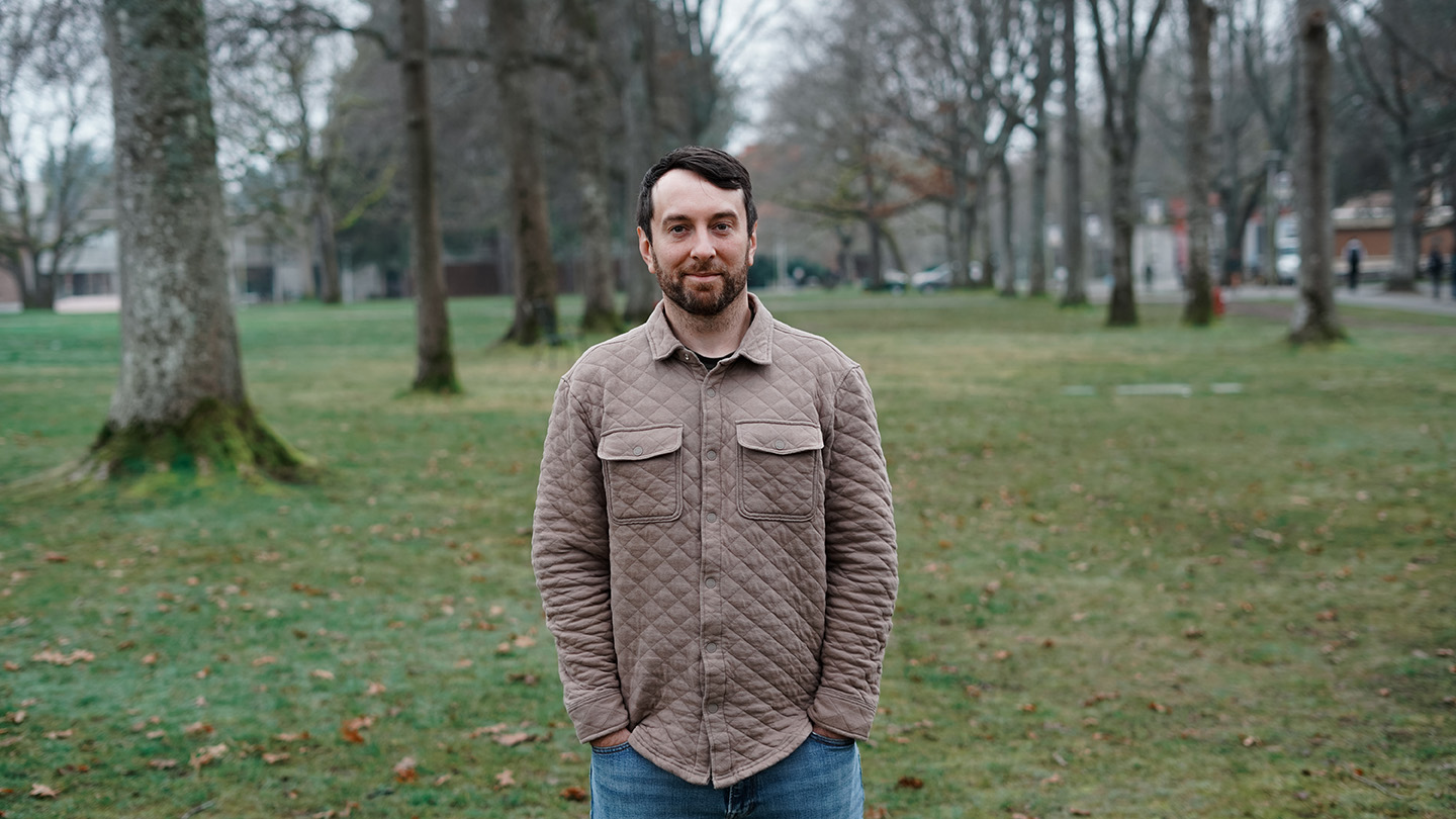 Bearded man wearing a tan quilted coat stands in the UVic quad, with green grass and bare trees in the background.