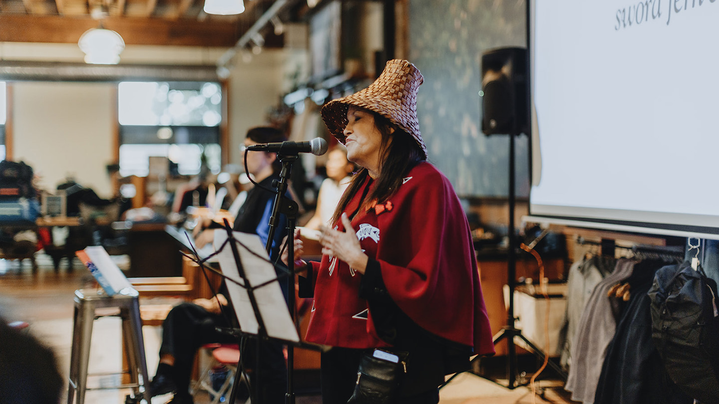 A woman wearing a traditional woven cedar hat speaks into a microphone on stage.
