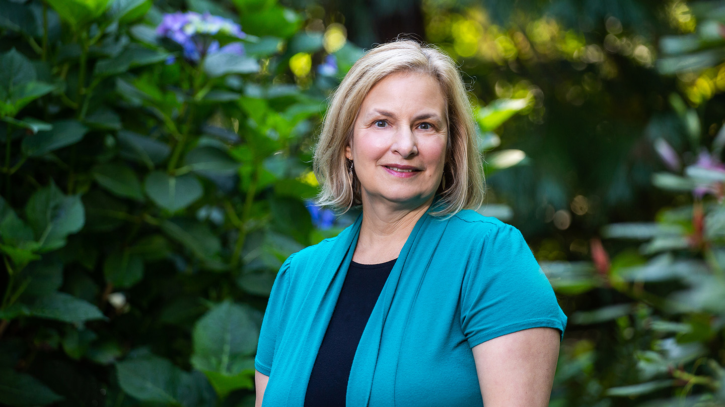 A blond woman in a teal shirt standing in front of a lush green background.