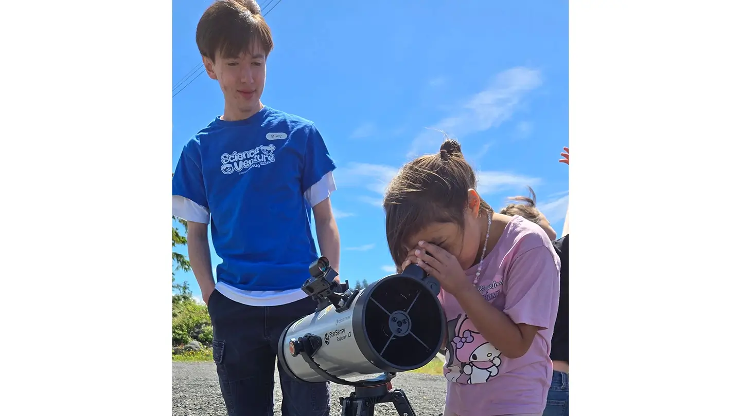 Riley Friesen stands next to a child as she looks into a telescope.