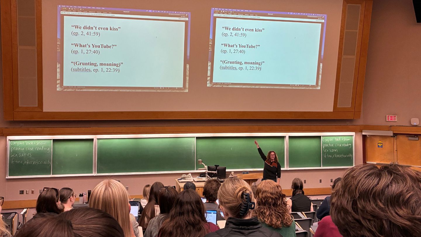 Janice Niemann presents to a full lecture theatre of students, pointing above her head to projected slides featuring iconic quotes from Heated Rivalry.