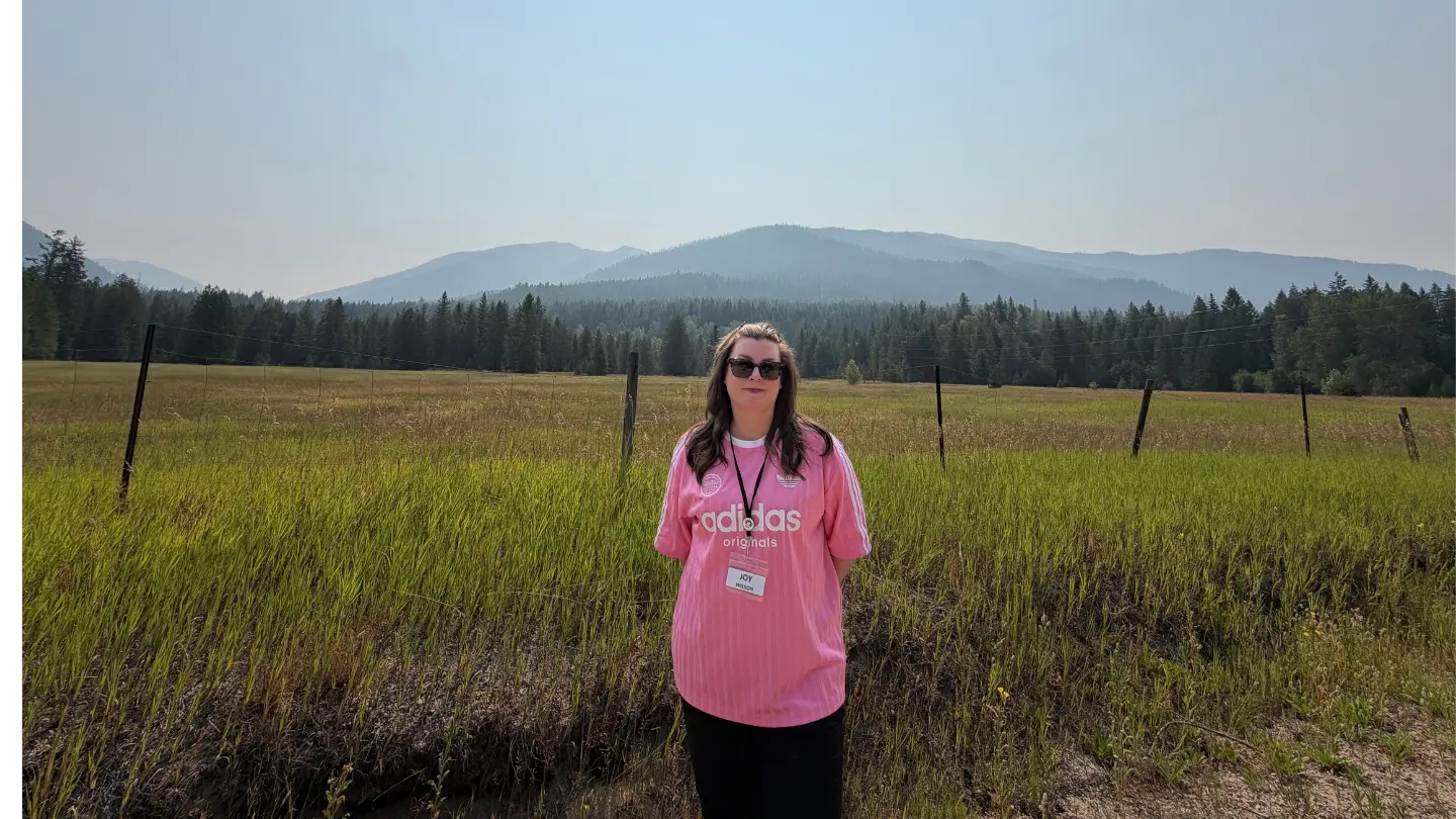 Joy Wilson wears a pink t-shirt and a lanyard. She is posing in front of a big, grassy field.